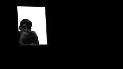 Young man looking thoughtfully through a window illuminated by bright light against dark surroundings.