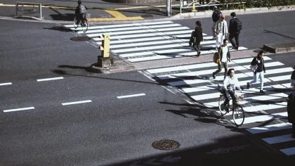 Pedestrians and cyclists cross a busy urban intersection with zebra crossings in a Japanese city scene.