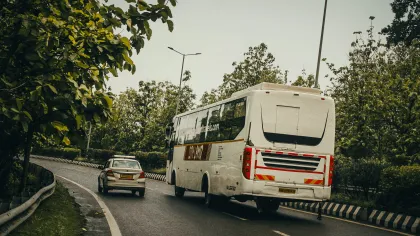 White passenger bus and car driving on curved road surrounded by green trees and street lamps