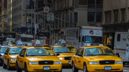Line of yellow taxis driving through a busy city street with trucks and buildings in the background