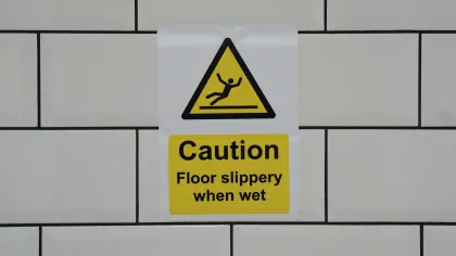 Yellow caution sign on white tiled wall warning floor slippery when wet with slipping figure icon.