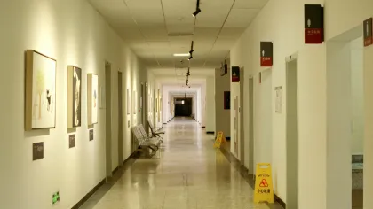 Long hospital corridor with artwork on the walls, chairs, restroom signs, and wet floor warning signs.
