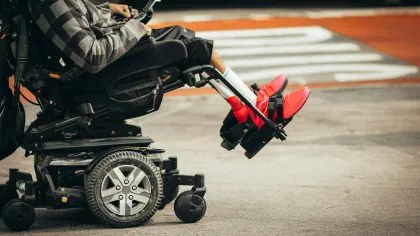 Person wearing red shoes and grey jacket sitting in an electric wheelchair on pavement near crosswalk.