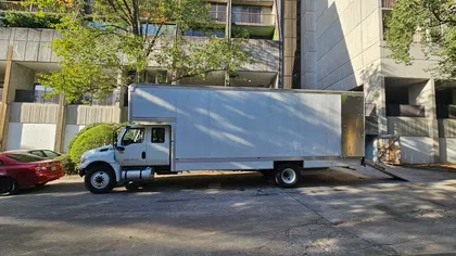 White delivery truck with open ramp parked on city street beside a red car during daytime.