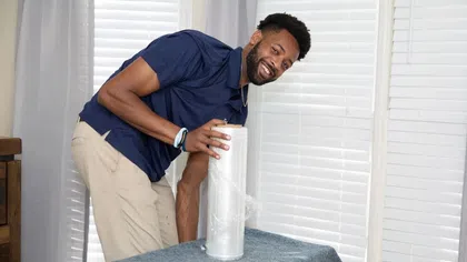 Smiling man preparing furniture with plastic wrap in a bright room with closed white blinds.