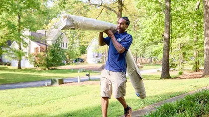 Man walking outside carrying a large rolled carpet over his shoulder on a sunny day in a suburban neighborhood