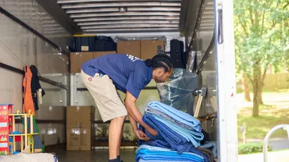 Mover stacking blue moving blankets inside a truck filled with boxes and furniture on a sunny day.