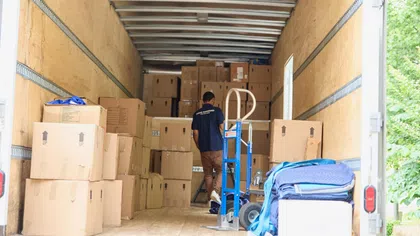 Worker unloading packed cardboard boxes inside a moving truck with a hand truck and moving blankets.