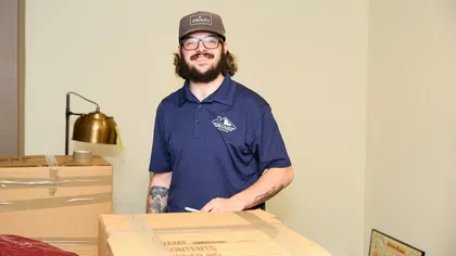 Smiling man with glasses and tattoos standing behind packed moving boxes in a room with beige walls.
