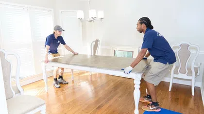 Two movers wearing uniforms and gloves lifting a wooden dining table in a bright room with hardwood floors.