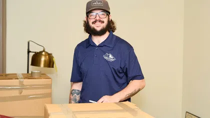 Smiling bearded man with glasses and cap packing boxes in a room with a lamp and framed pictures.