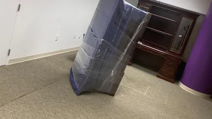 A wrapped mattress leaning against a carpeted office room wall with a wooden cabinet and purple pillar.