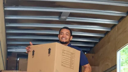 Smiling man carrying a large cardboard box inside a moving truck filled with packed boxes under natural light.