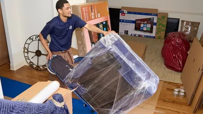 Mover transporting wrapped furniture on a dolly inside a home surrounded by packed boxes and protective coverings.