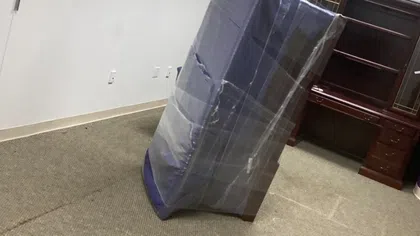 Plastic-wrapped mattress standing on carpeted floor in office room with wooden desk and purple pillar.