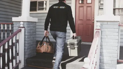 Man wearing Detect and Defeat jacket carrying tool bags walks up porch steps to house entrance