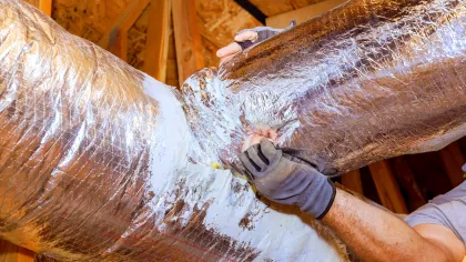 Worker insulating and sealing ductwork with foil insulation in a wooden attic space for HVAC system.