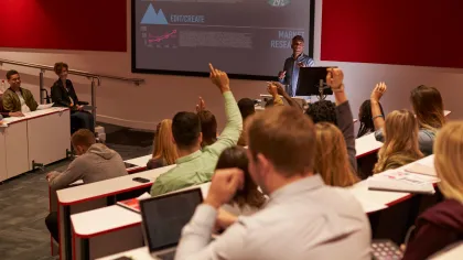 Students raising hands in a modern lecture hall while a professor presents data on a screen.