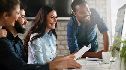 Four diverse coworkers smiling and collaborating at a computer in a bright modern office environment