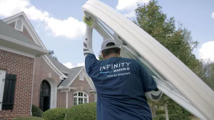 Worker in Infinity by Marvin shirt carrying a large window frame outside a brick house on a sunny day.