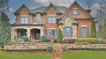 Large two-story brick and stone suburban house with manicured lawn, shrubs, and stone steps leading to the entrance.