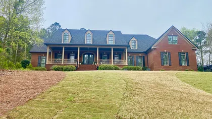 Large brick house with black shutters, dormer windows, and a covered porch, surrounded by green and dry lawn.