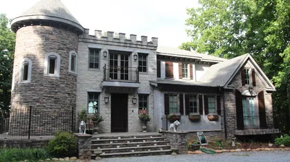 Stone house with turret, balcony, shuttered windows, and landscaped front stairs amid greenery on a sunny day