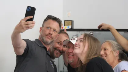Group of five people smiling and taking a selfie indoors with a smartphone.