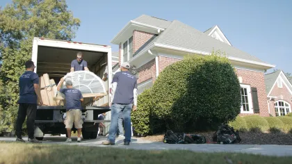 Workers unloading a large window frame from a truck outside a brick house on a sunny day