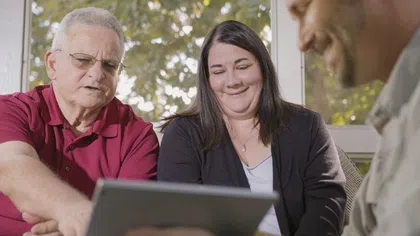 Three adults smiling and looking at a tablet together in a bright room with large windows.