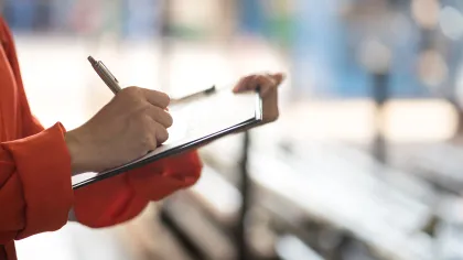 Person in orange sleeve writing on clipboard with blurred background of benches and sunlight.