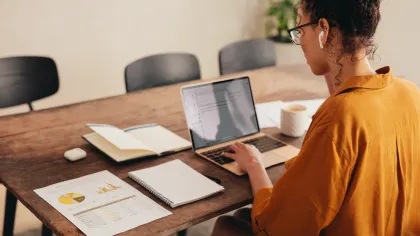 Woman in orange shirt working on laptop with documents and notebooks on wooden table in office.