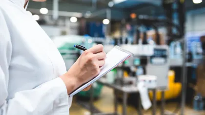 Person in lab coat writing notes in notepad inside a modern industrial or laboratory setting.