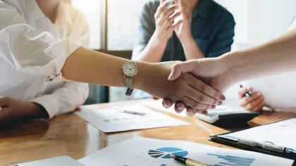 Two professionals shaking hands over business charts and laptops in a meeting room with natural light.