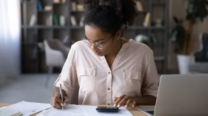 Woman wearing glasses calculating finances with a calculator and documents at a wooden table with laptop.