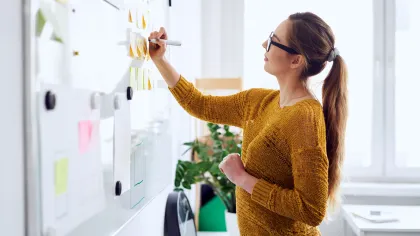 Woman in sweater writing on sticky notes on a whiteboard in a bright office space with plants.