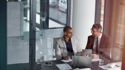 Two businessmen in suits discussing work at a table with laptops, documents, and tablets in a modern office.