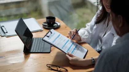 Two people discussing a business report on a clipboard at a wooden table with a tablet, eyeglasses, and coffee cup.