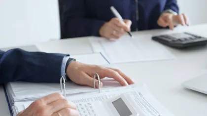 Two people calculating finances with a white calculator and documents at a desk in an office setting.