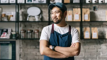 Smiling male barista in blue apron and cap stands with crossed arms in modern coffee shop interior.