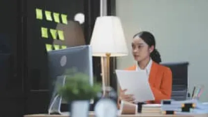 Professional woman in orange blazer working at desk with computer, papers, and office supplies in modern workspace.