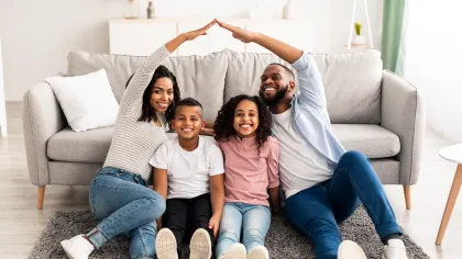 Happy family of four sitting on carpet in living room, parents making a roof shape with their hands above children.
