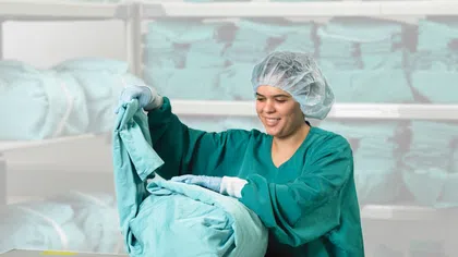 Healthcare worker in scrubs folding clean medical linens in a storage room with stacks of blue sheets.
