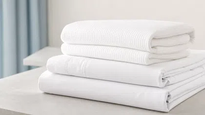 Neatly folded white bed sheets and textured towels stacked on a light gray surface near blue curtains.