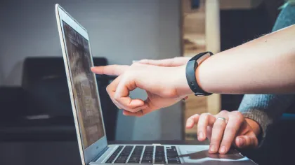 Person pointing at laptop screen while another types on keyboard in a casual indoor setting.