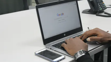 Hands typing on an HP laptop with Google search page open, smartphone placed beside on a white office desk.