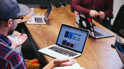 Team meeting with laptops on wooden table focusing on a man presenting app design wireframes on screen.