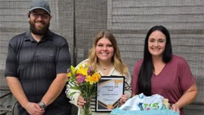 Three smiling people stand in front of a wooden wall, one holding flowers and certificate, another with a gift basket.