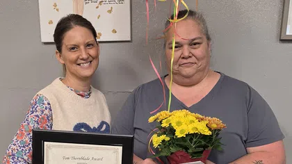 Two women pose indoors with a certificate, flowers, and balloons during an award presentation event.