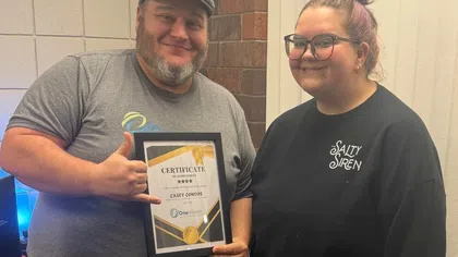 Two smiling people posing indoors with a certificate of achievement in a black frame